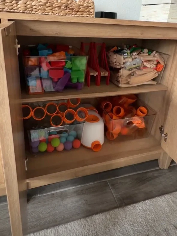 Clear storage bins of blocks, wooden train tracks and orange tubes on two shelves inside a wooden cabinet