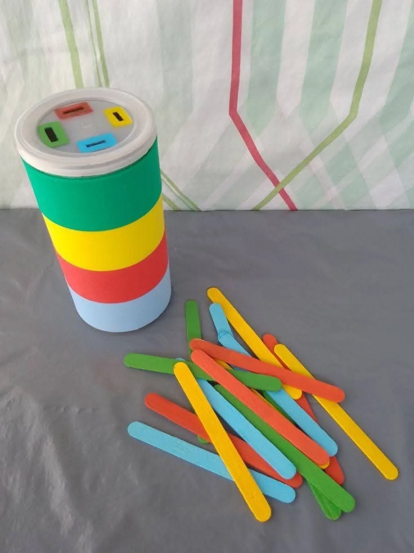 A canister wrapped in green, yellow, red and blue bands with four colored slots in its lid, beside scattered craft sticks