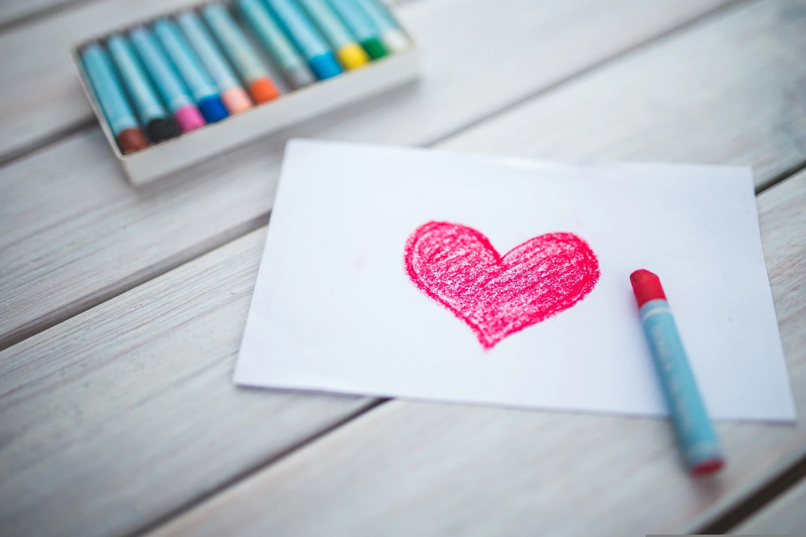 Open box of oil crayons on a white wooden table beside a sheet of paper with a red heart drawn on it