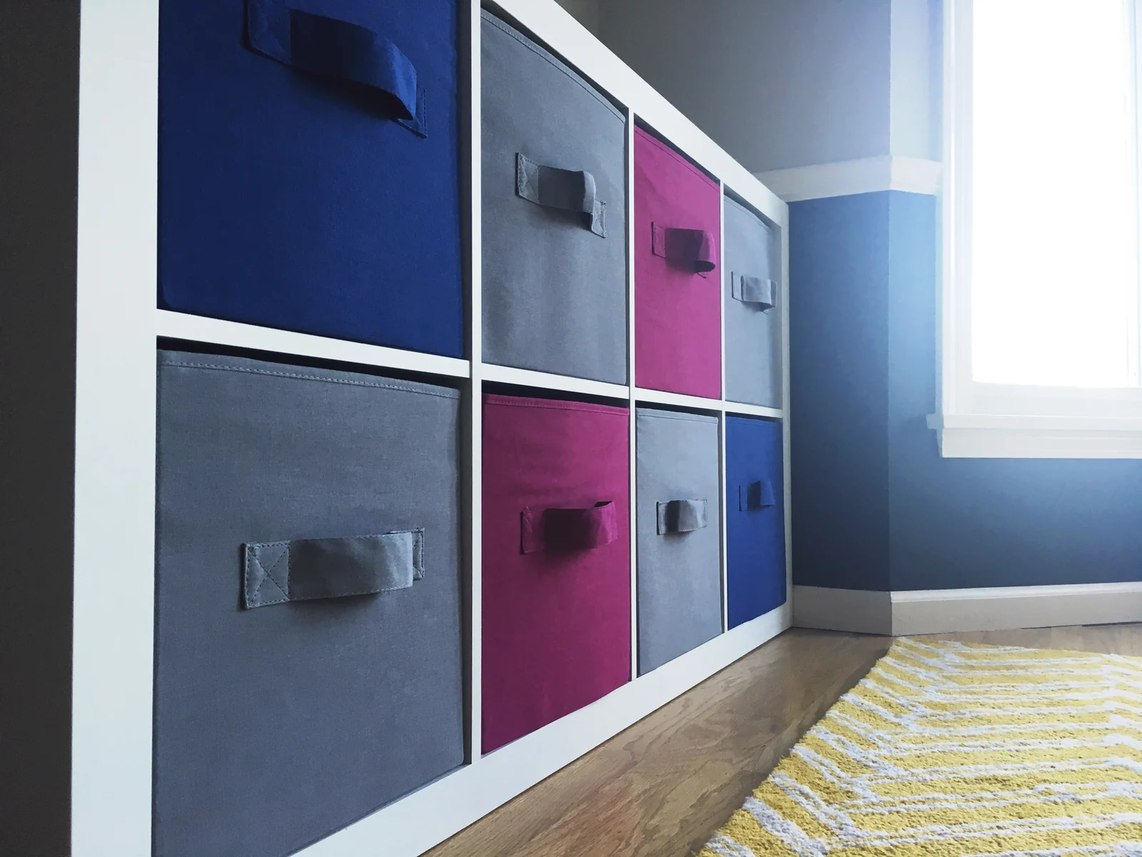 A white cube shelf with closed fabric storage bins in blue, gray and magenta, beside a window and a yellow rug