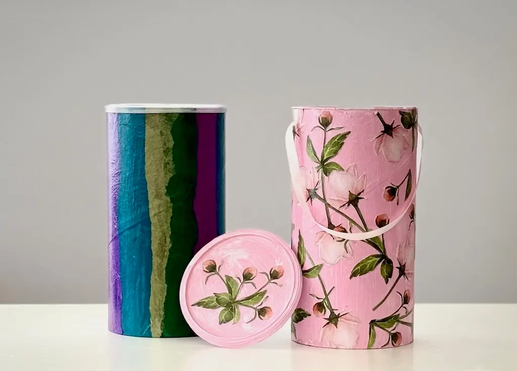 Two paper-covered cardboard canisters, one striped in purple, teal and green, one pink with flowers, with a loose lid
