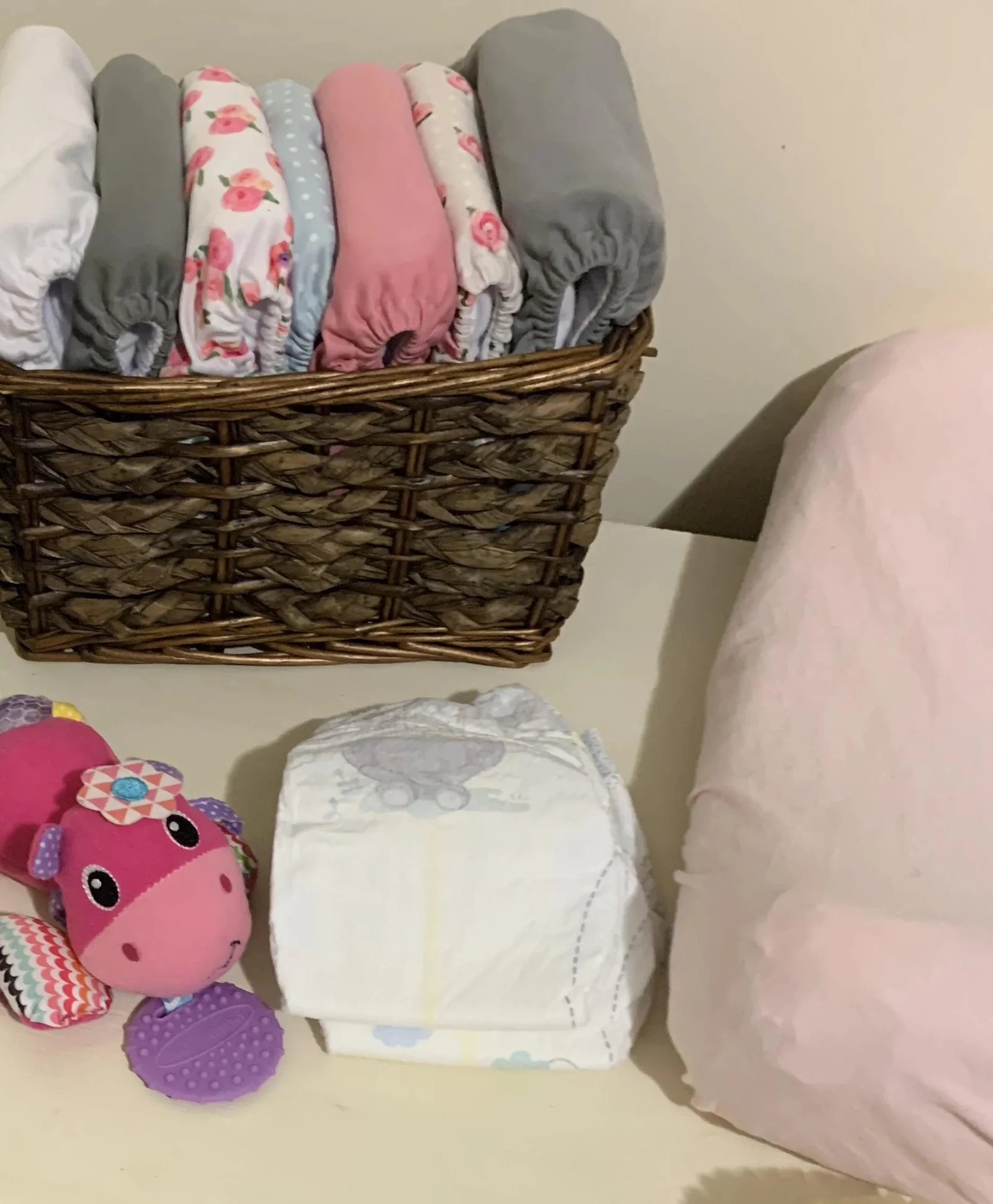 Wicker basket of rolled cloth diaper covers on a changing table next to a newborn diaper and a plush baby toy