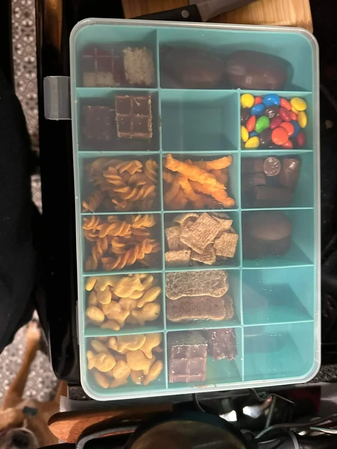 Clear-lidded snack box with teal compartments holding crackers, chocolate and candy, photographed from above