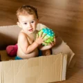 A baby sits in a cardboard box on a wood floor holding a textured green and blue ball: DIY Montessori activities