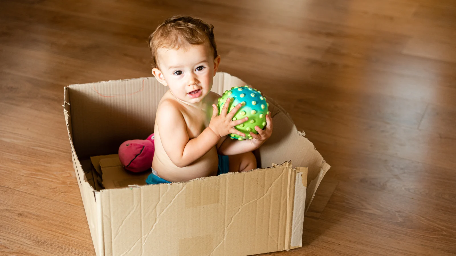 A baby sits in a cardboard box on a wood floor holding a textured green and blue ball: DIY Montessori activities