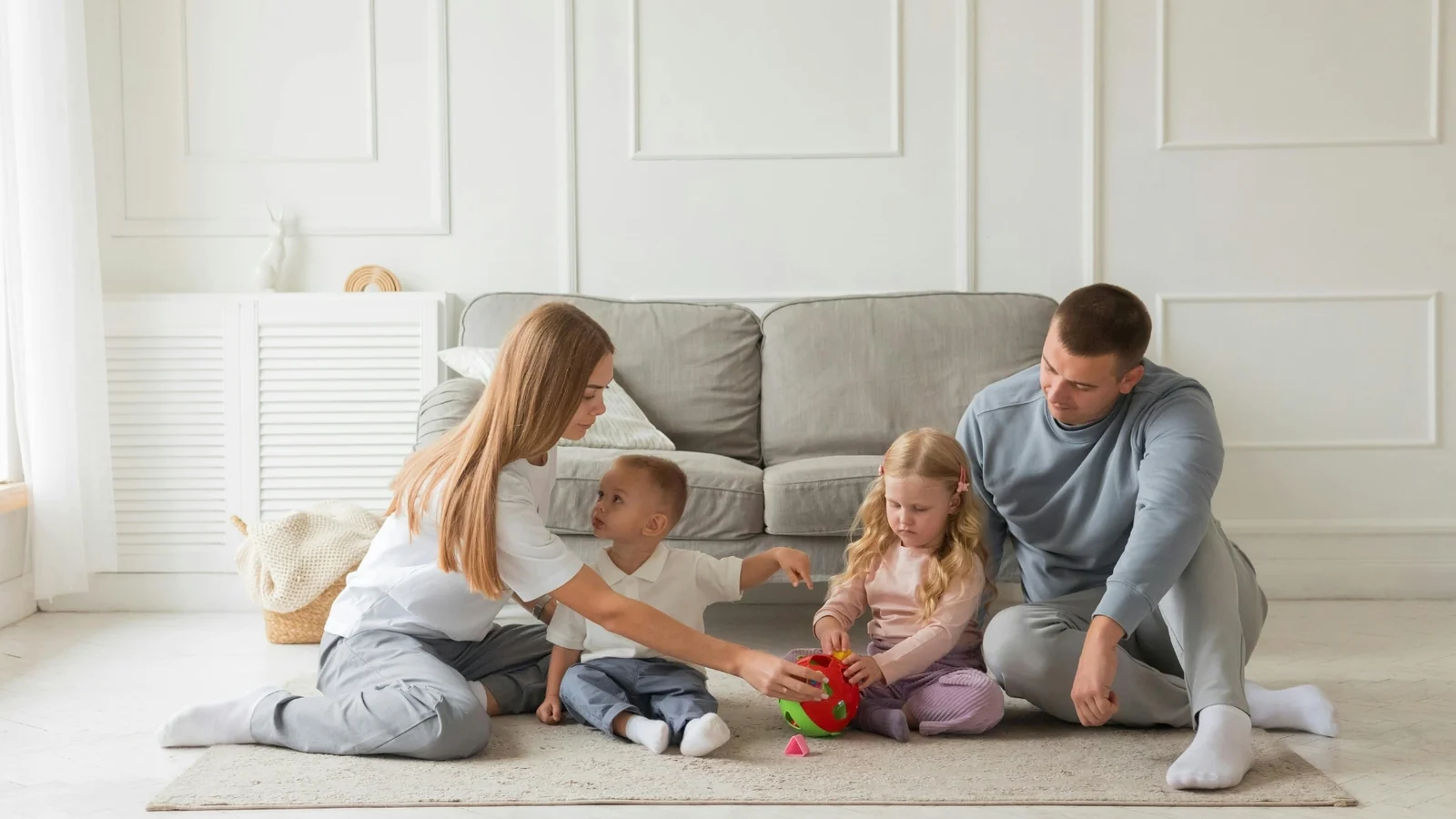 A family of four plays together on a rug in a bright living room: a Montessori playroom corner