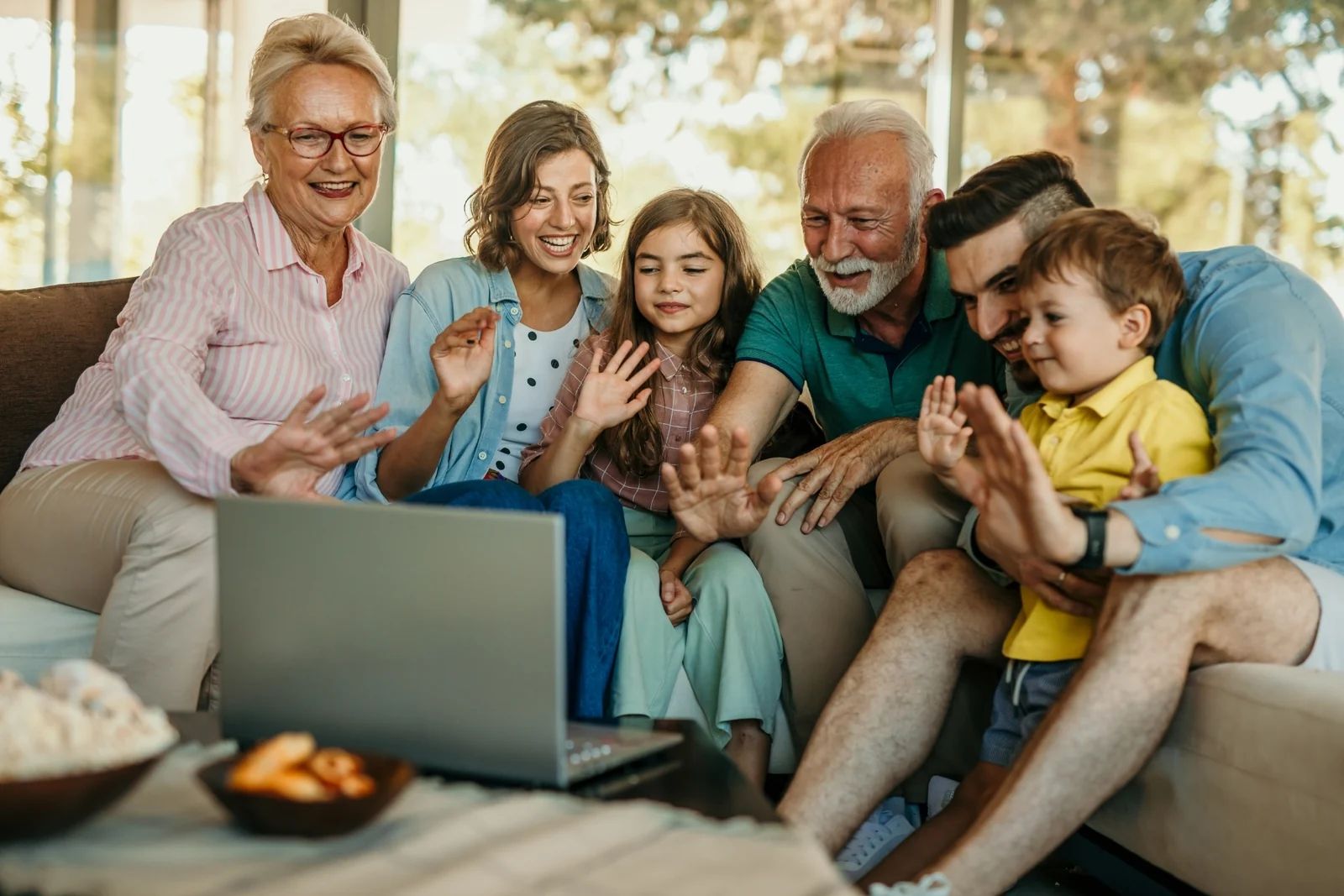 Three generations of a family on a couch waving at a laptop together for a family video chat, a toddler among them