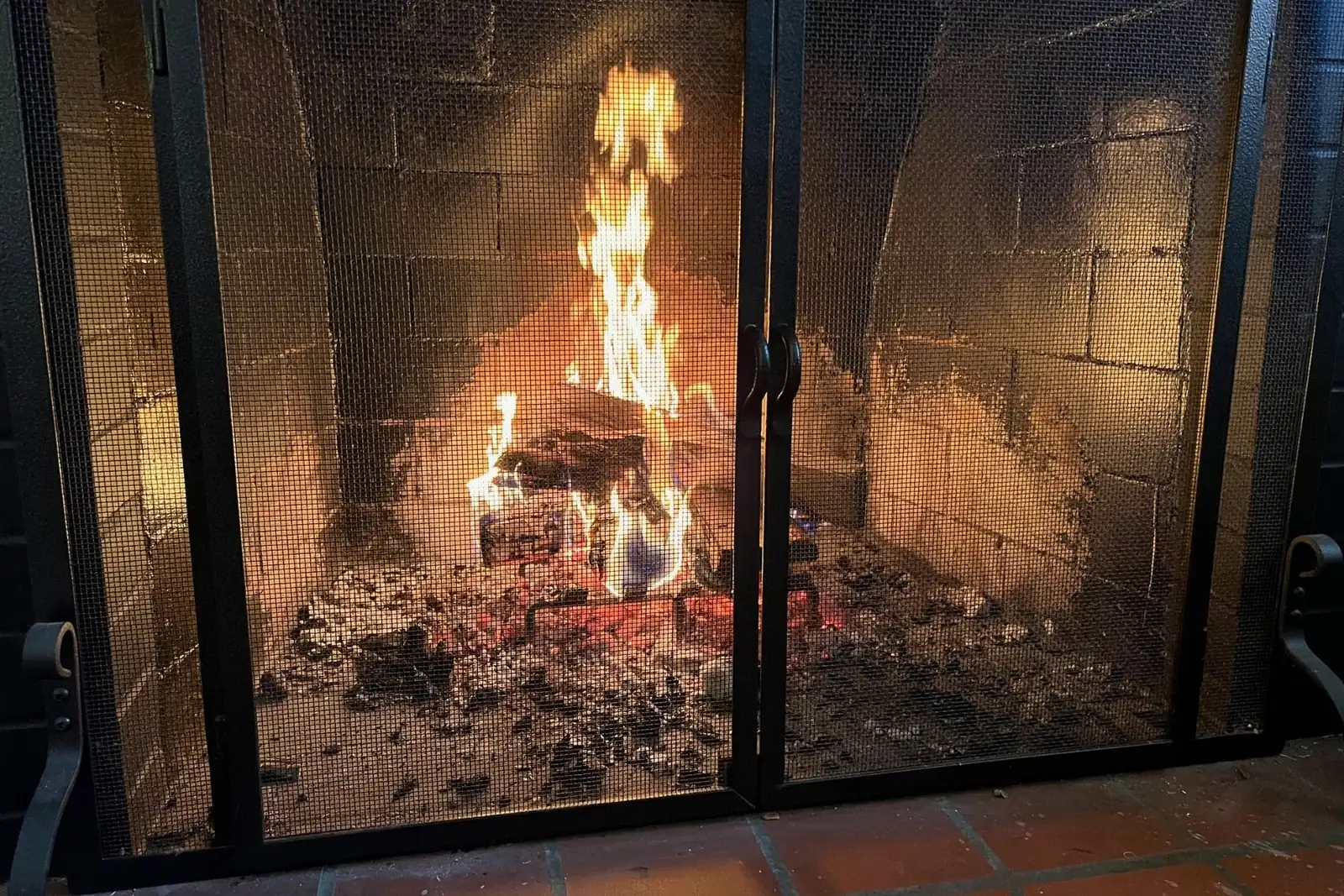 A wood fire burns in a brick fireplace behind a black metal mesh screen with two doors