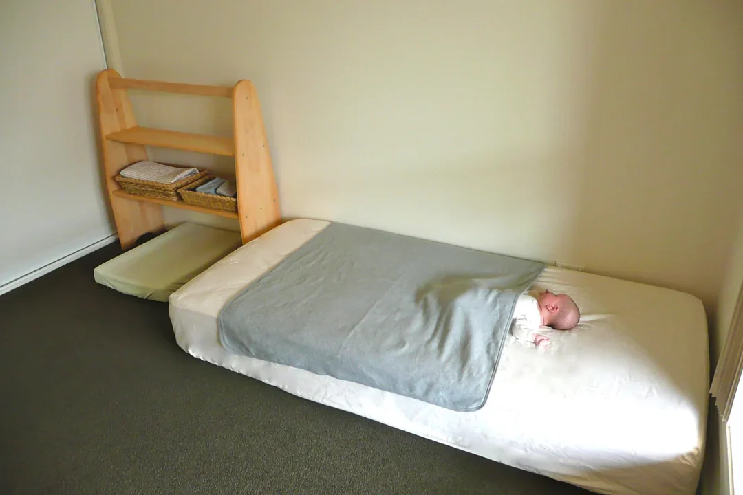 A baby lying under a grey blanket on a mattress set straight on the carpeted floor, a small wooden shelf beside it