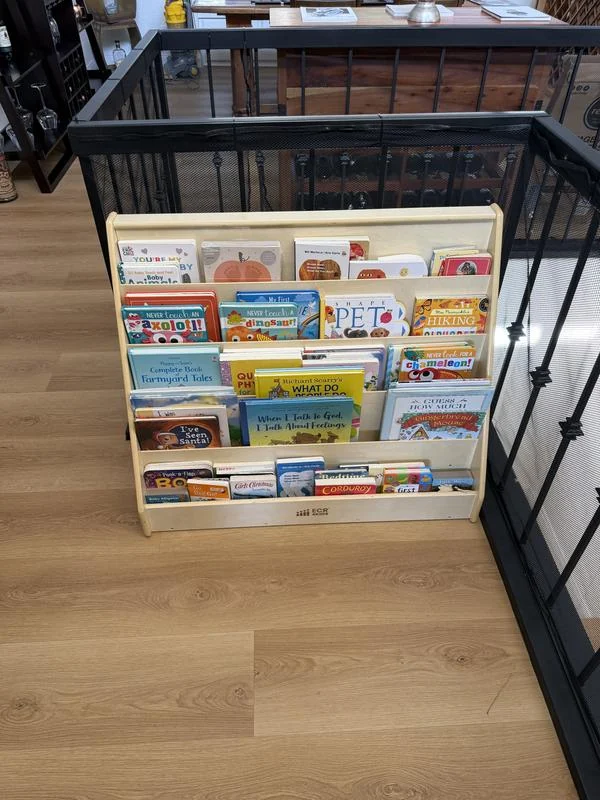 A five-tier wooden front-facing bookshelf full of picture books, standing on a wood floor beside a black railing