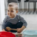 A toddler squats on a kitchen floor with a wooden spoon beside a red colander and a pale blue bowl for free montessori activities.
