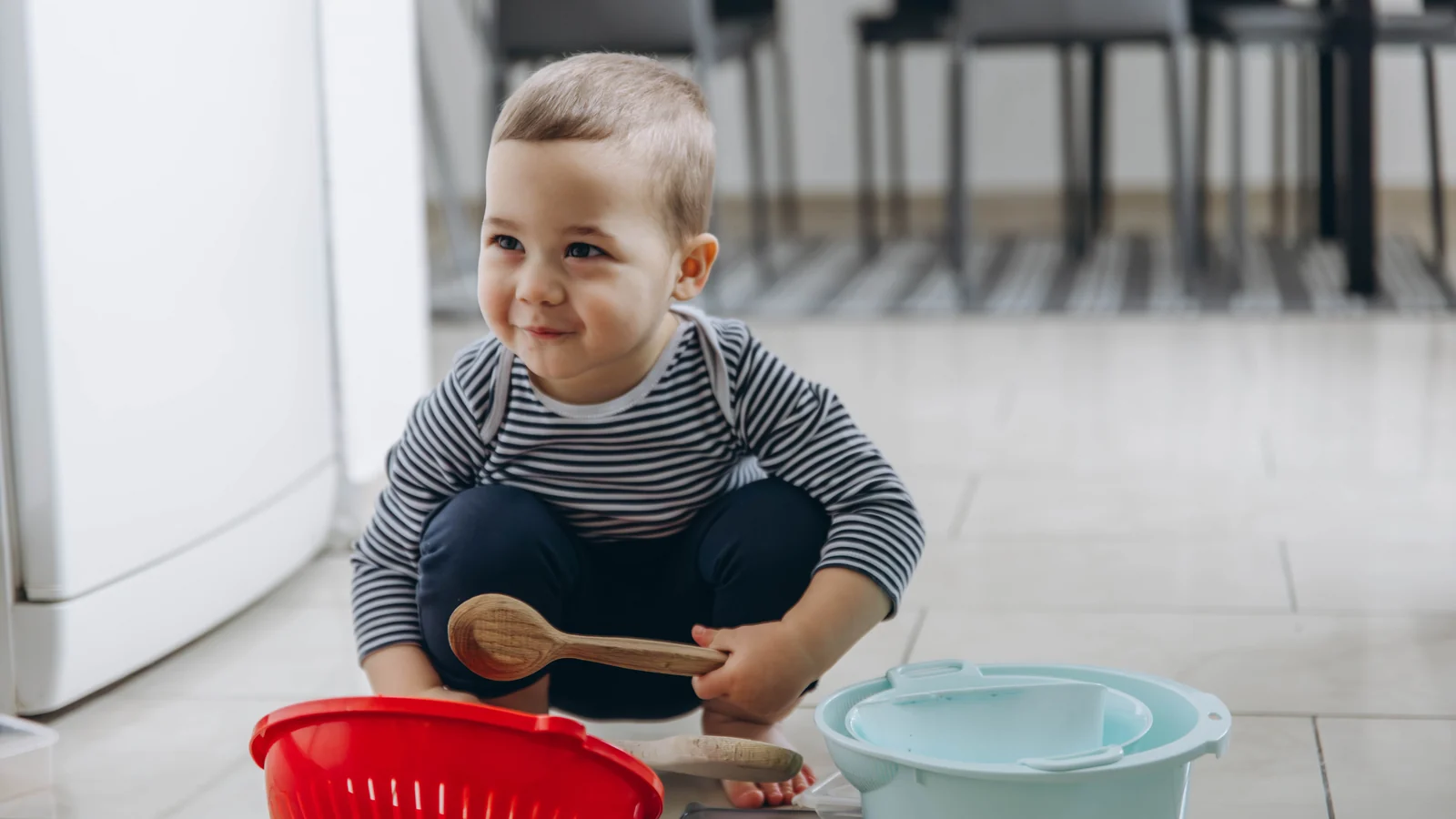 A toddler squats on a kitchen floor with a wooden spoon beside a red colander and a pale blue bowl for free montessori activities.