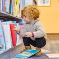 A toddler crouches at a low library shelf, picking a picture book alone: the choice a front facing bookshelf makes easy