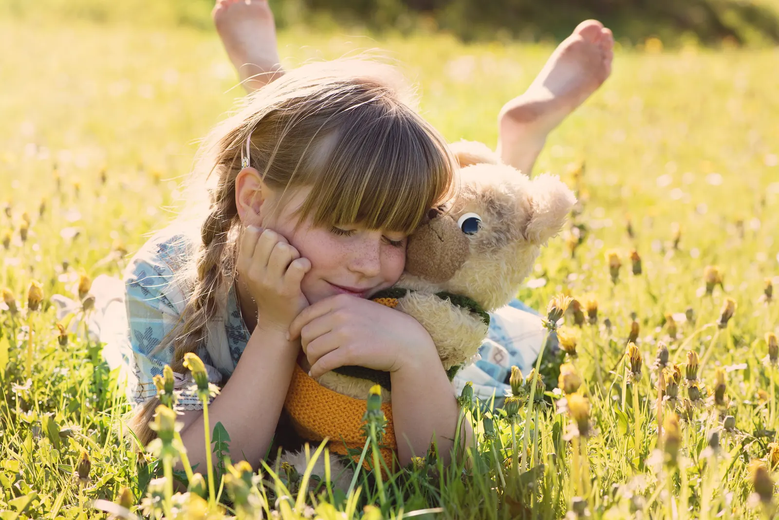 Girl lying in a meadow with her cheek resting on a plush teddy bear she is hugging