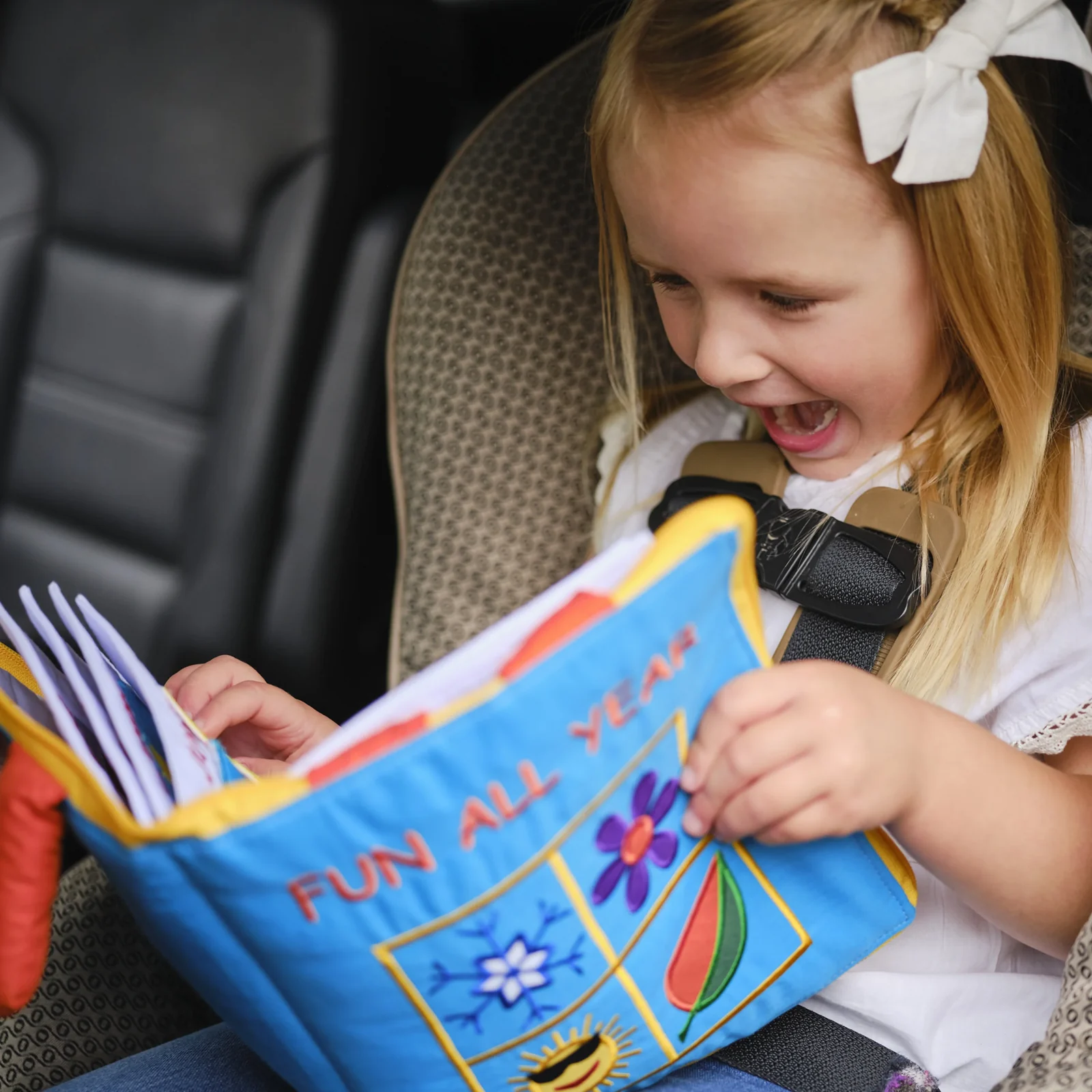 Girl in a car seat laughing as she opens a blue cloth book with a flower, a leaf and a sun on the cover