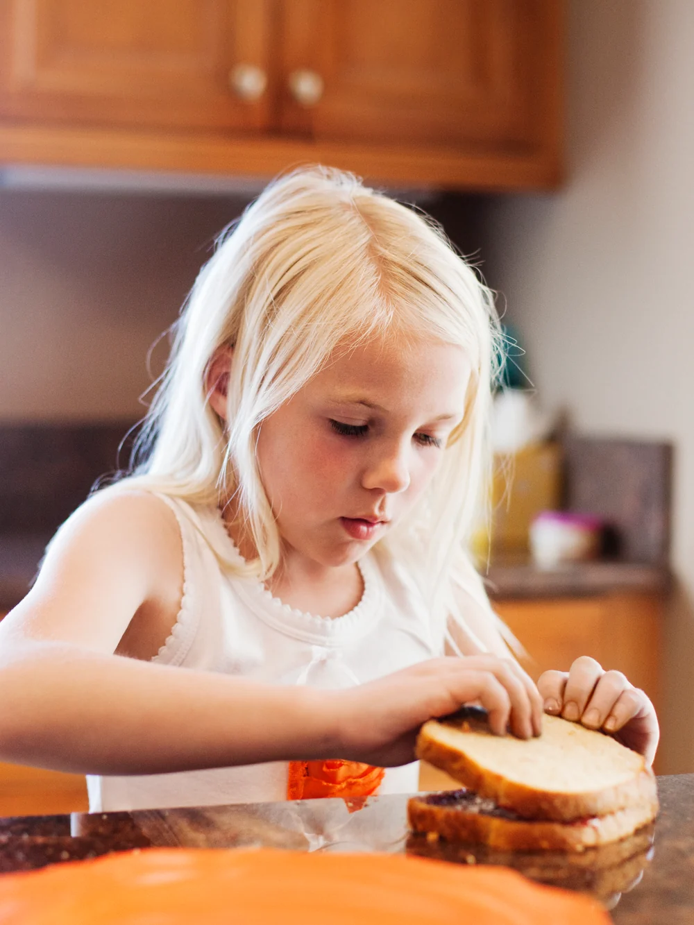 Girl making a jam sandwich at a kitchen counter, pressing two slices of bread together, a jar of jam beside her