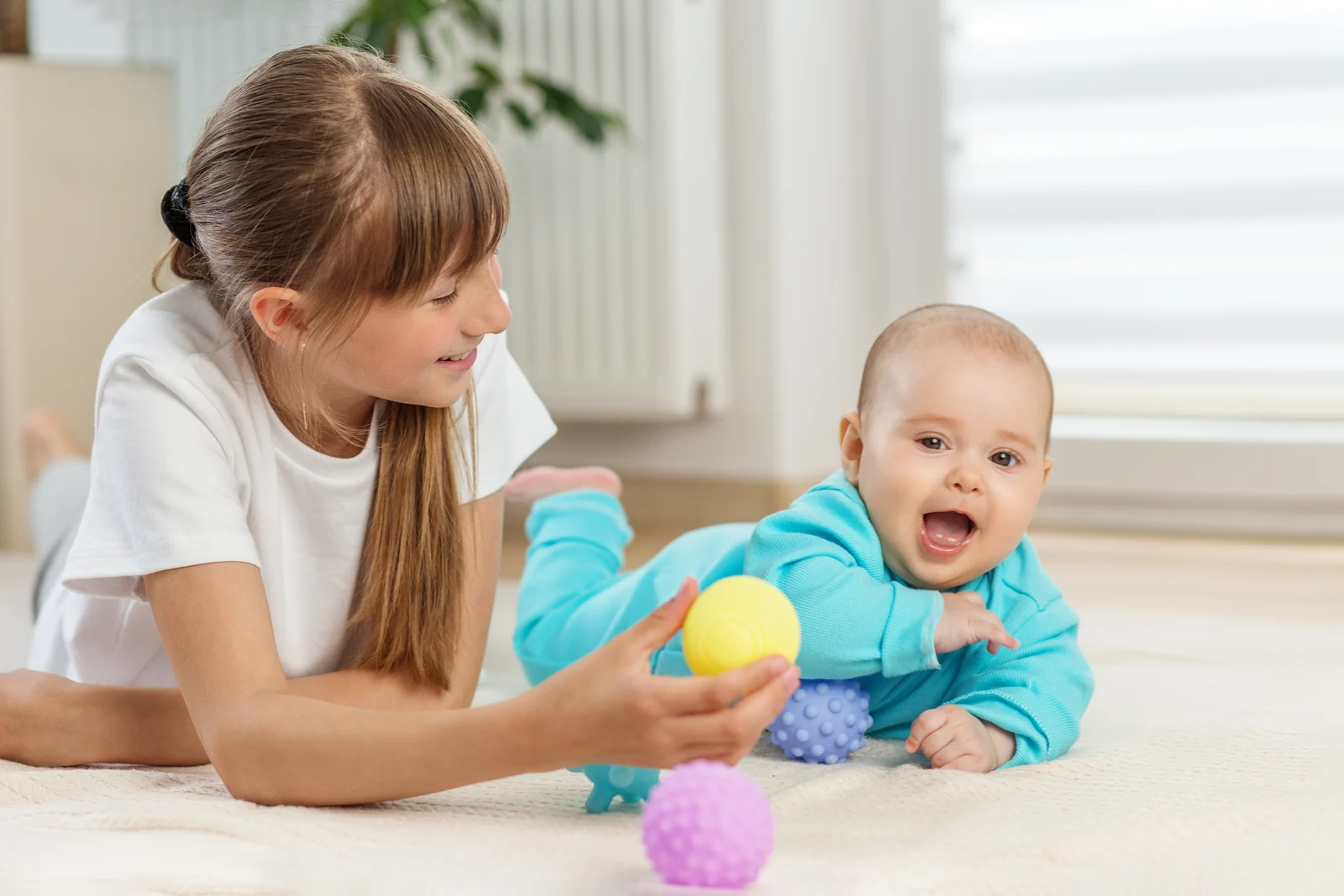 Girl lying on a rug holding a textured yellow ball toward a smiling baby lying on their tummy