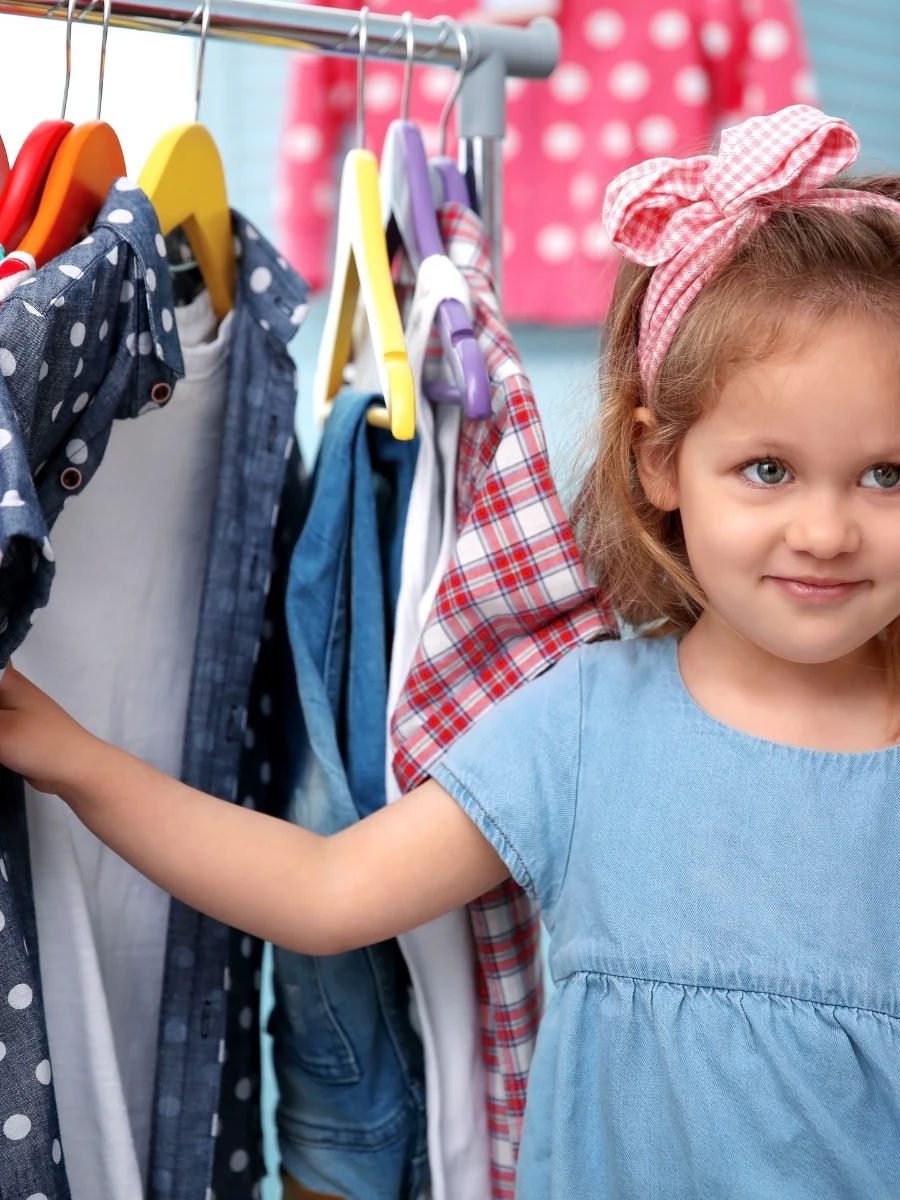 A smiling girl reaches toward a rack of colorful children's clothes on hangers