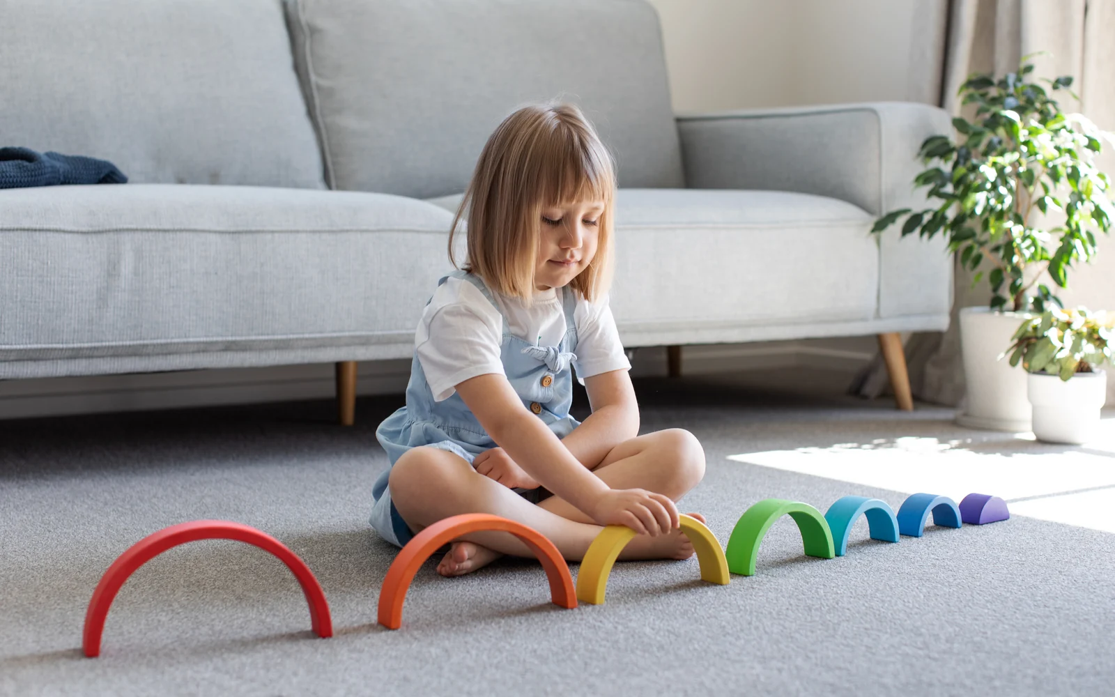 A young girl sits on a gray rug beside a row of wooden rainbow arches set out in a line