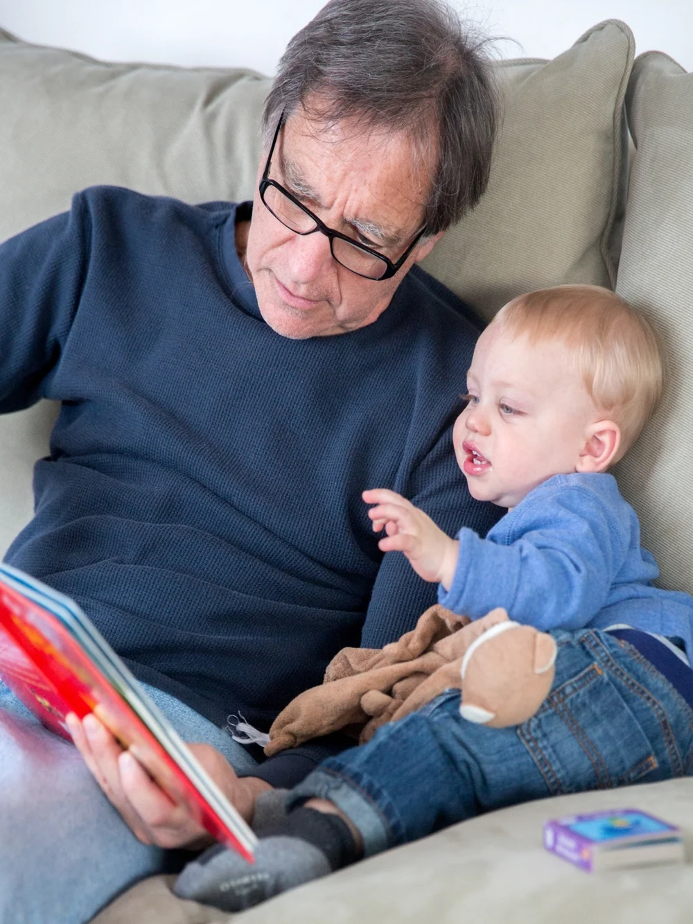 Grandfather with glasses reading a colorful picture book to a baby sitting beside him on the couch