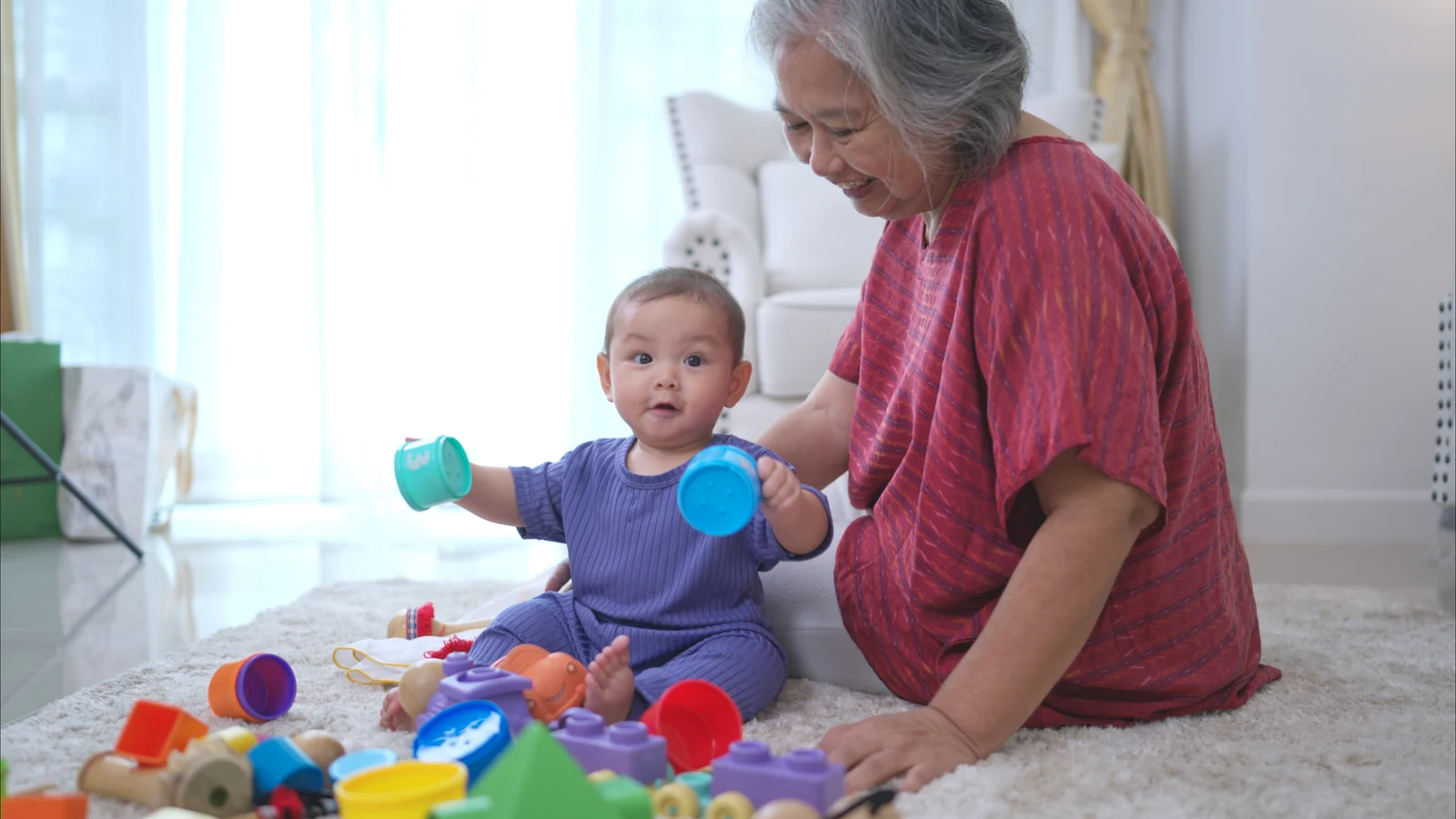 Grandmother sitting on a rug beside her baby grandchild, who holds two toy cups from a pile of colorful toys