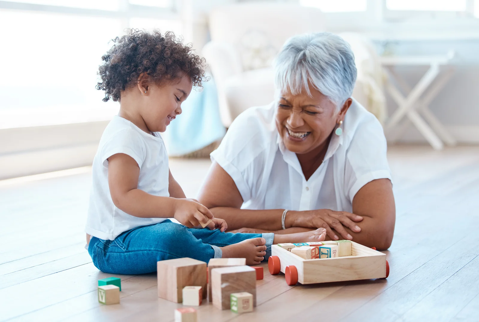 Grandmother laughing on the floor beside a toddler, with wooden blocks and a wooden wagon between them