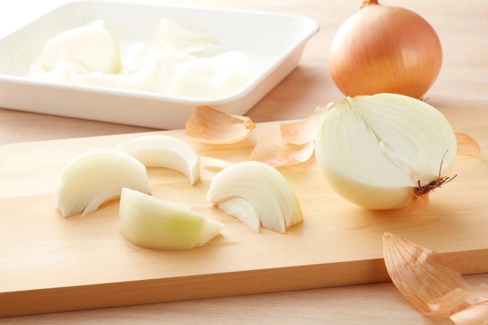 Onion wedges and a cut half onion on a light wooden cutting board, with a whole onion and a tray of slices behind