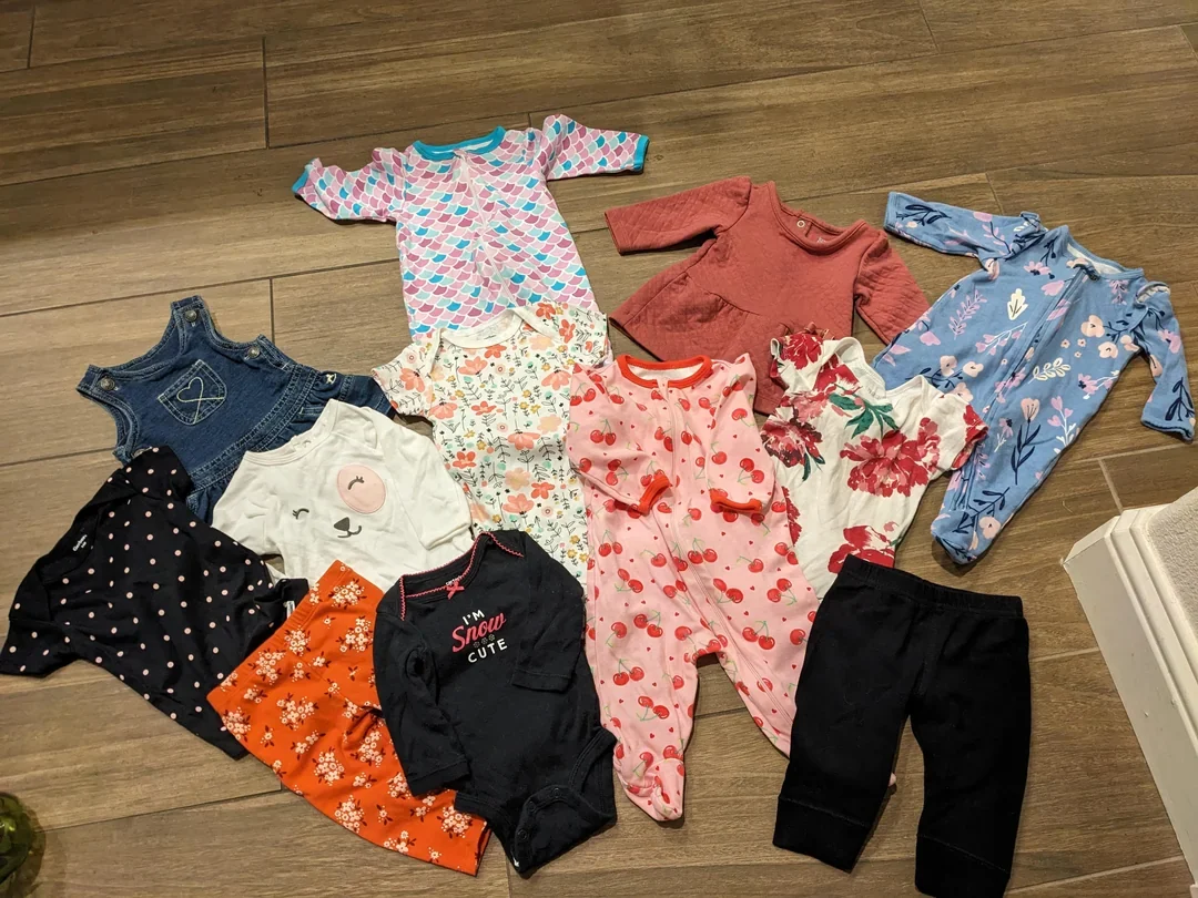 Baby clothes spread out on a wood-look floor: sleepsuits, bodysuits, overalls, a skirt and leggings