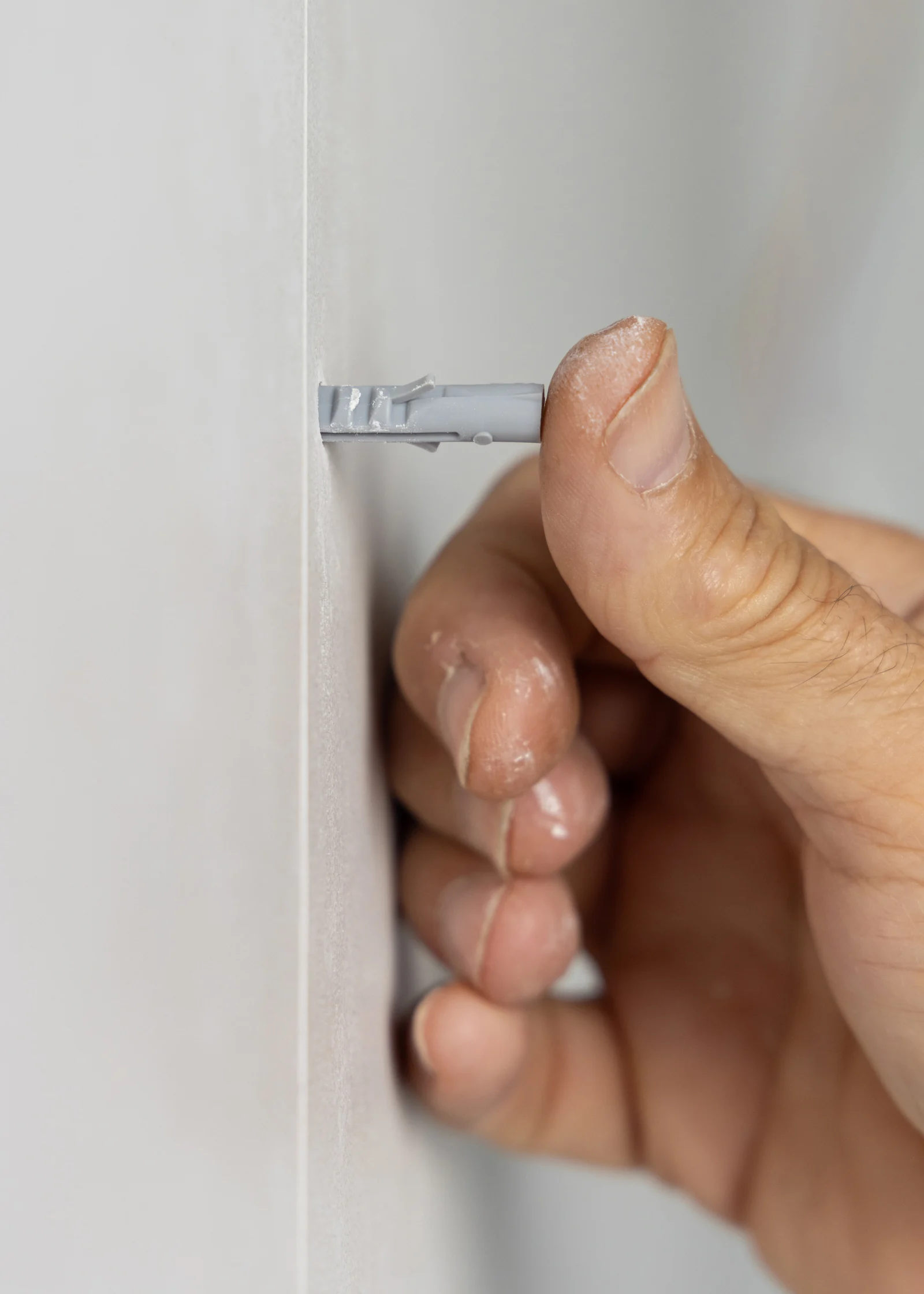 A hand presses a gray plastic wall plug into a drilled hole in a white wall