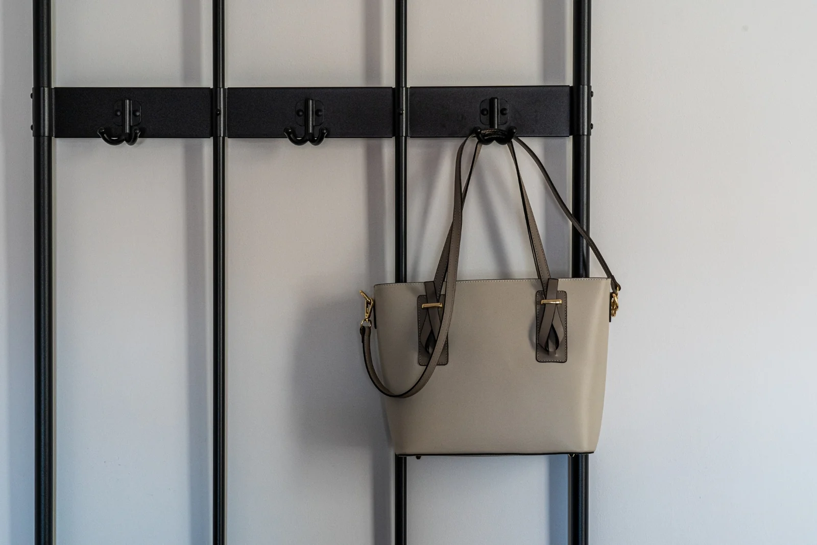 A gray handbag hanging from a black metal wall hook on a pale wall