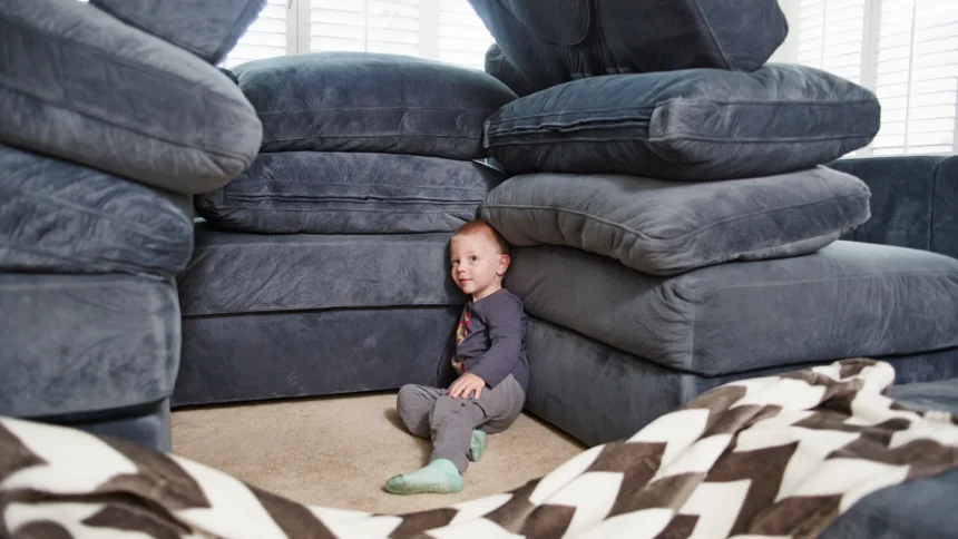 Toddler sitting beside a fort built from stacked sofa cushions in a living room: indoor activities for toddlers