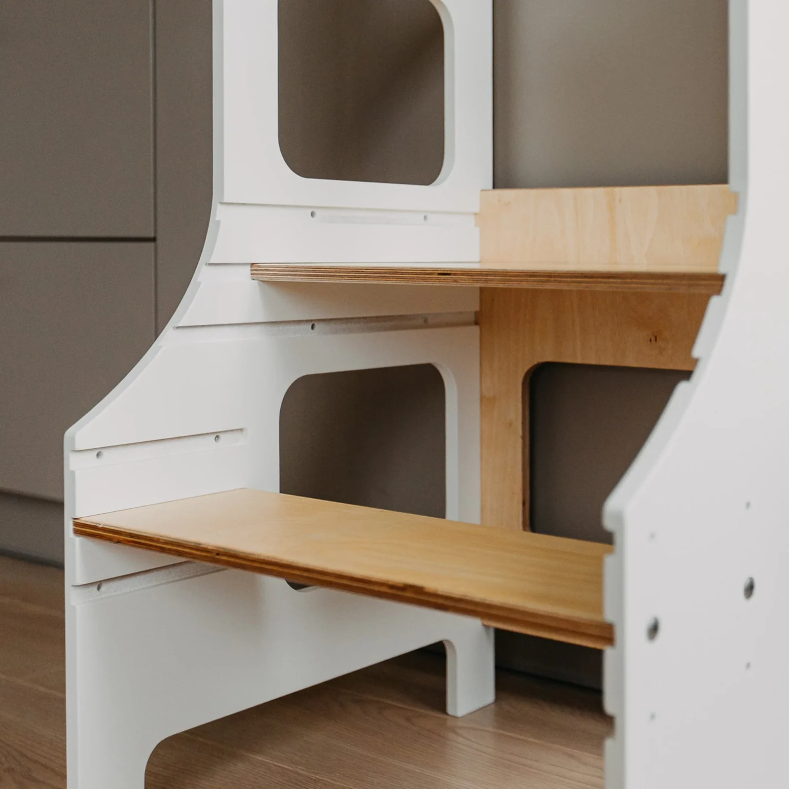 Close-up of a white learning tower with plywood platforms, rounded cut-out openings and platform-height slots