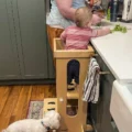 A toddler standing in a wooden learning tower at the kitchen counter, an adult right beside them working with fresh greens