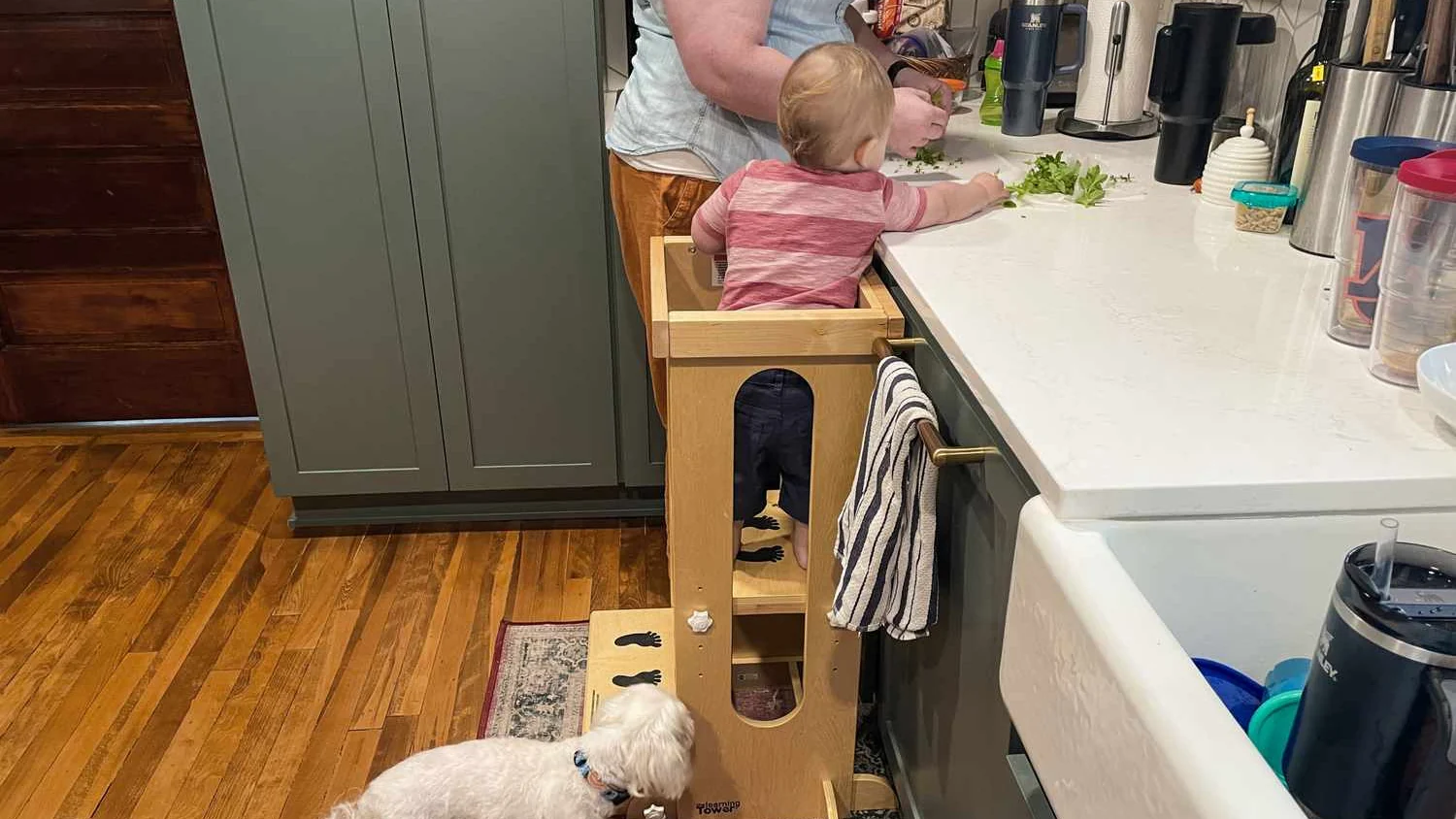 A toddler standing in a wooden learning tower at the kitchen counter, an adult right beside them working with fresh greens
