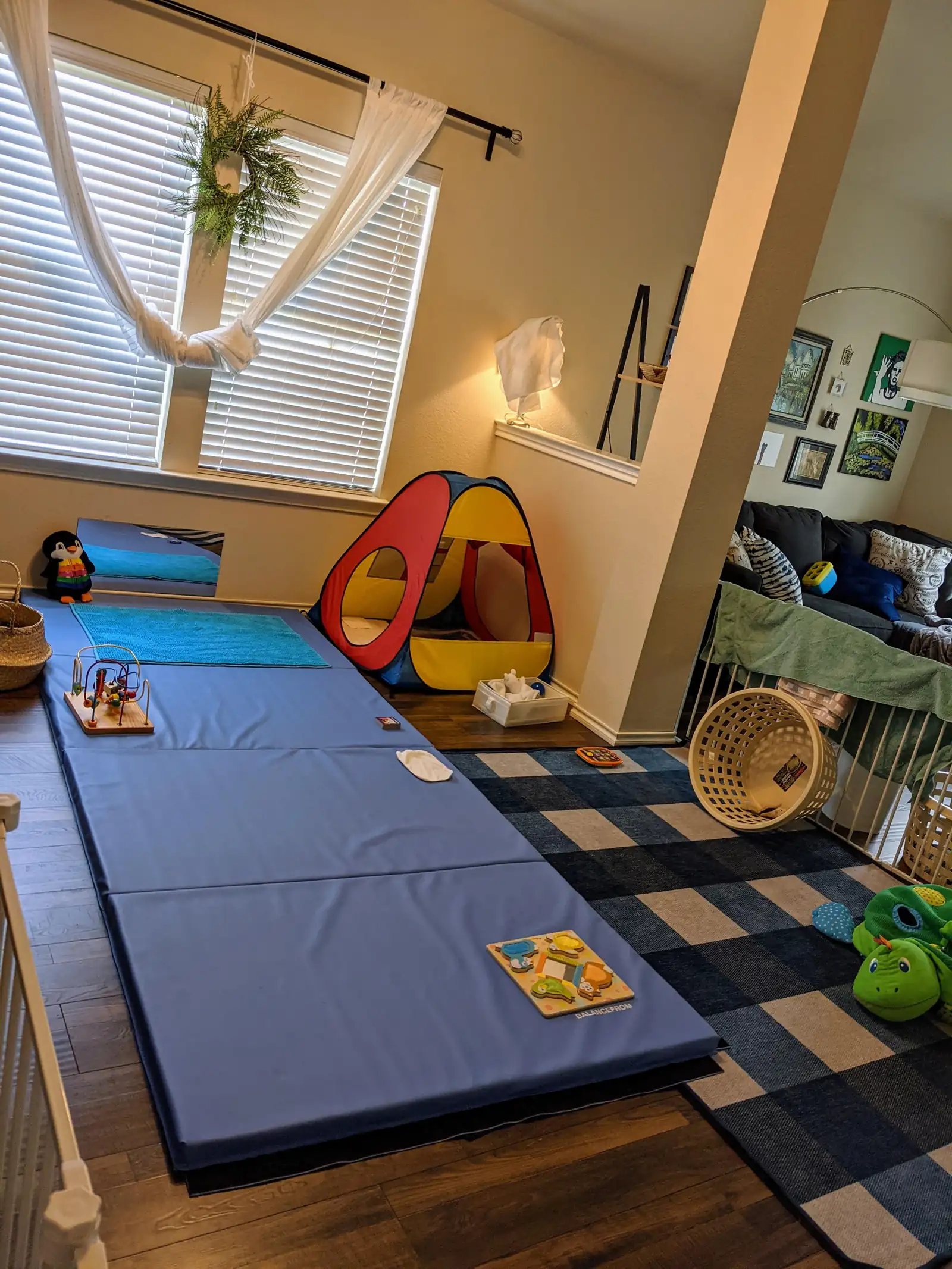 A living-room play corner with a blue folding floor mat, a pop-up tent, a wooden bead maze and a plaid rug by a window