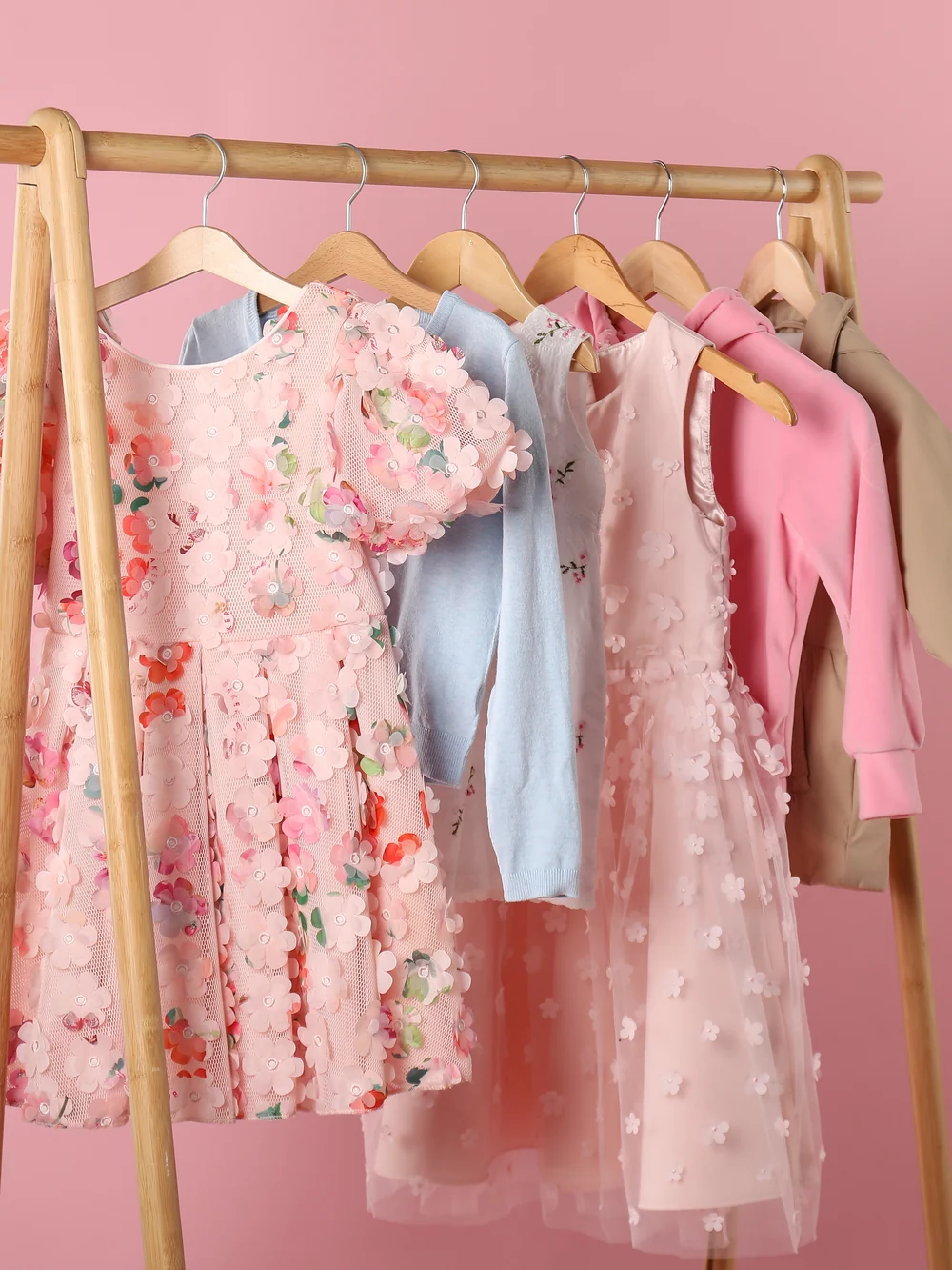 Small wooden clothes rack with a few children's dresses and jackets on wooden hangers against a pink wall