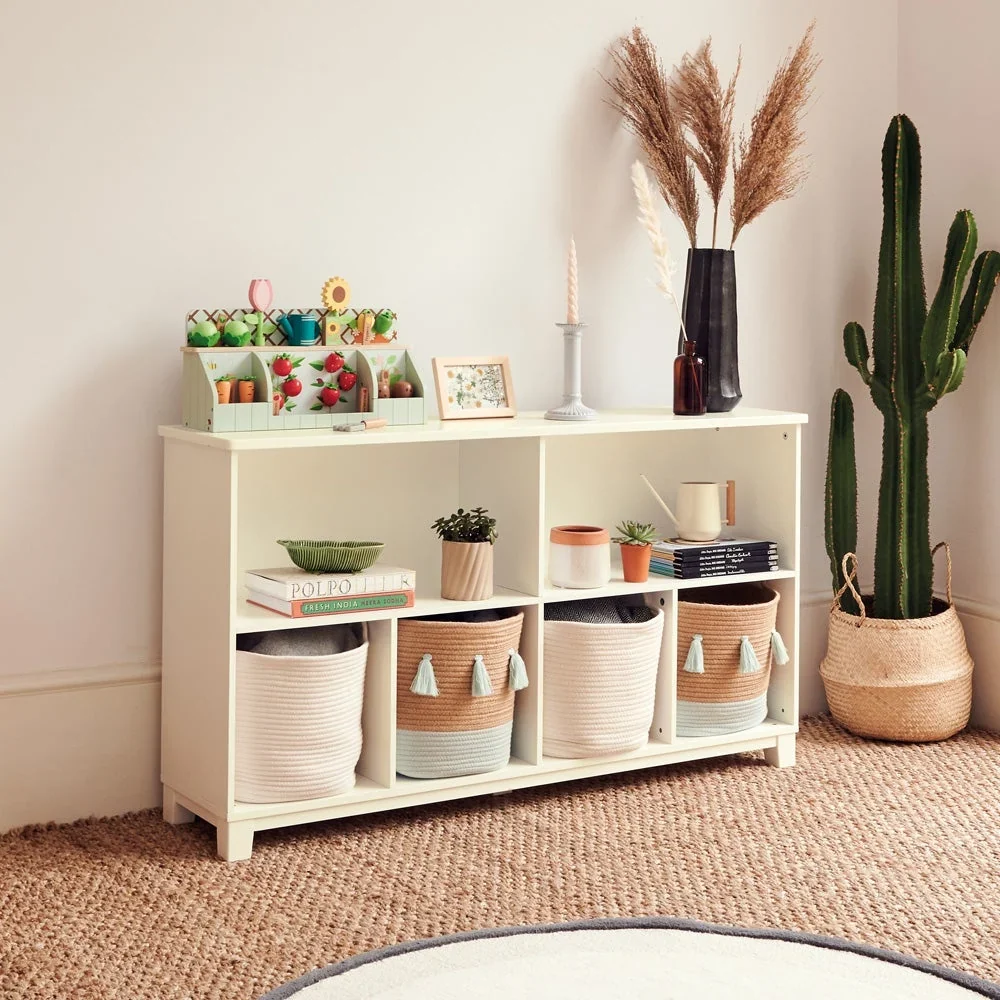 A low cream shelf with woven rope baskets, books and small plants, with a wooden garden toy set on top, beside a jute rug