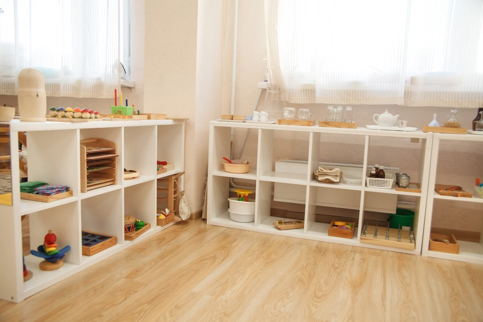 Low white open shelves in a bright room hold Montessori toys in baskets and trays