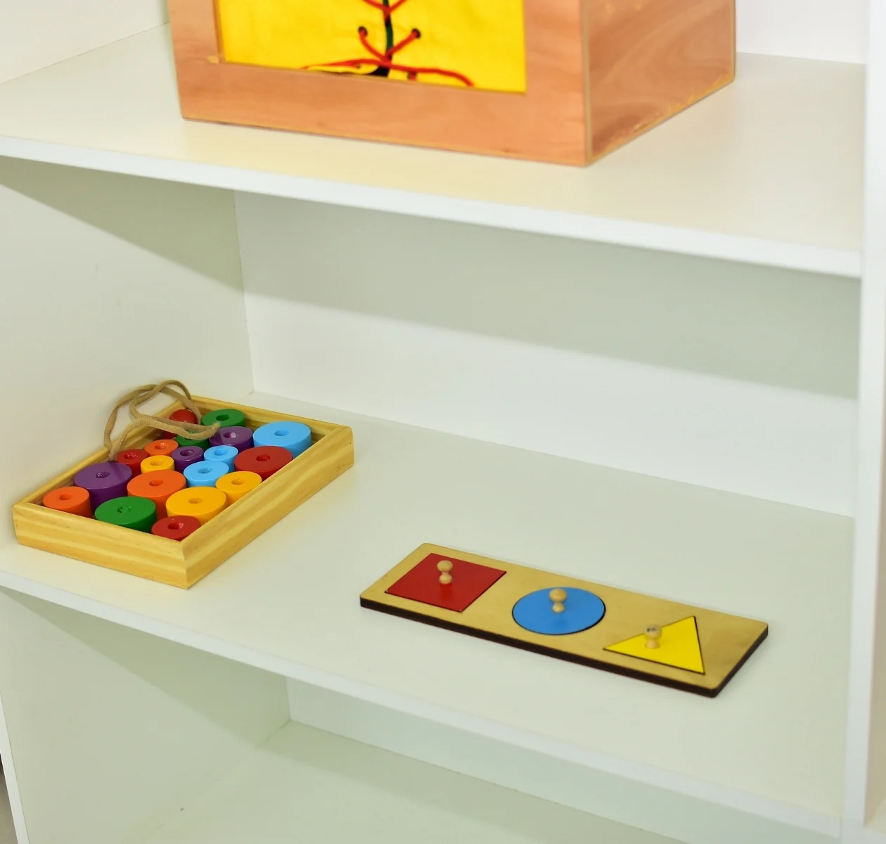 White open shelf with two wooden toys spaced apart: a knobbed shape puzzle and a tray of colorful wooden pieces