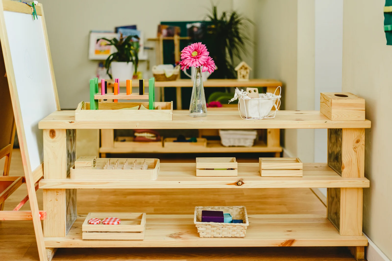 Low wooden shelf with a few trays and baskets and a glass vase of flowers on top, beside an easel in a bright room