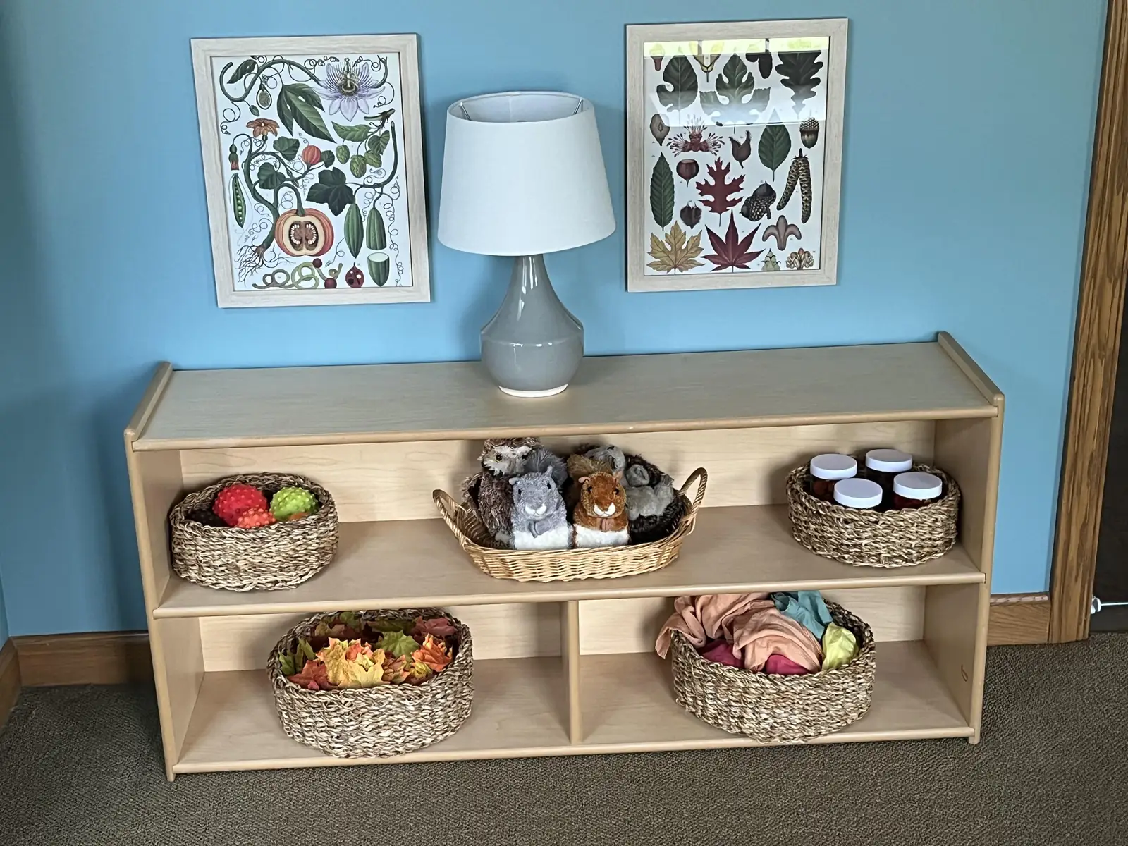 Low wooden shelf with five woven baskets of balls, plush animals, jars and scarves under a lamp and two framed prints
