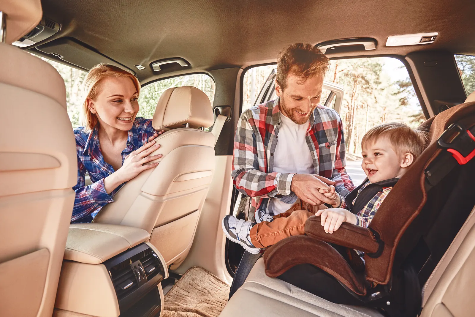 Man holding a laughing toddler's hand in a car seat while a smiling woman looks on, rear car door open