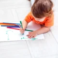 Toddler lying on the floor drawing with a colored pencil on a sheet of paper, a simple Montessori art activity