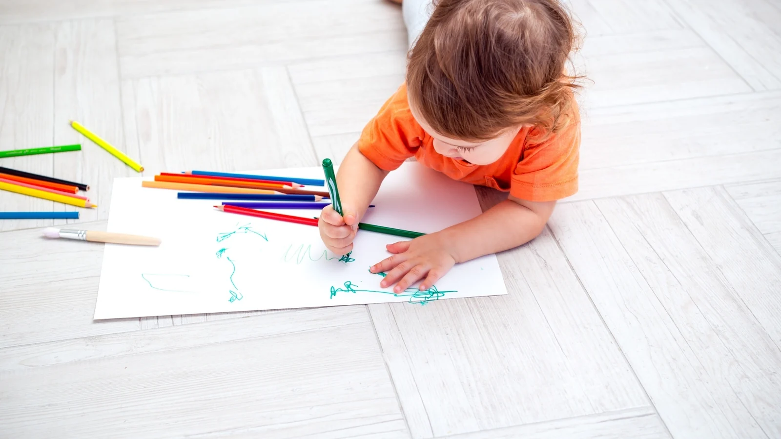 Toddler lying on the floor drawing with a colored pencil on a sheet of paper, a simple Montessori art activity