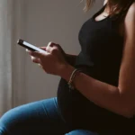 Pregnant parent sitting by a window, reading about Montessori criticism on a phone