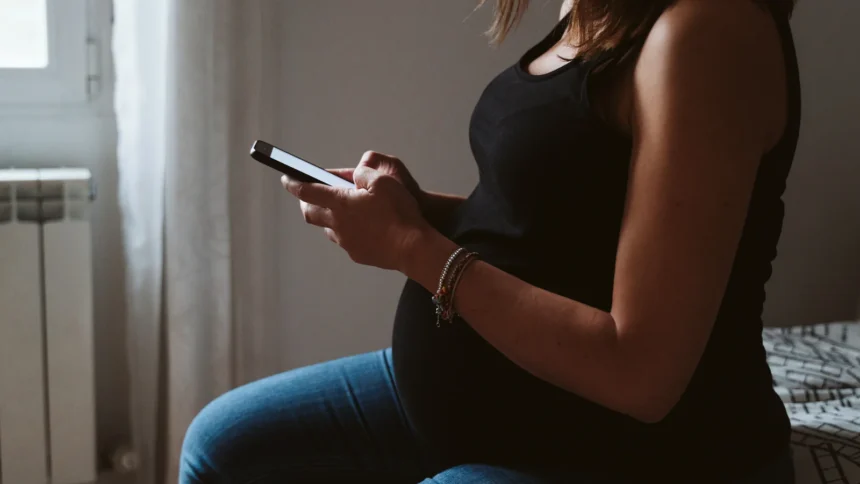 Pregnant parent sitting by a window, reading about Montessori criticism on a phone