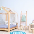 A low wooden house-frame bed in a bright child's room: a Montessori floor bed setup with a small table and stools