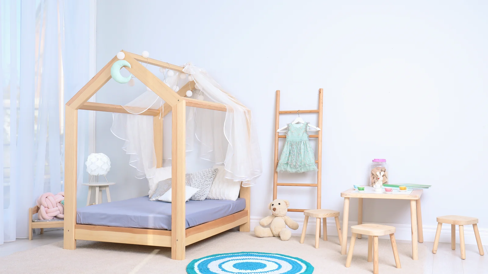 A low wooden house-frame bed in a bright child's room: a Montessori floor bed setup with a small table and stools