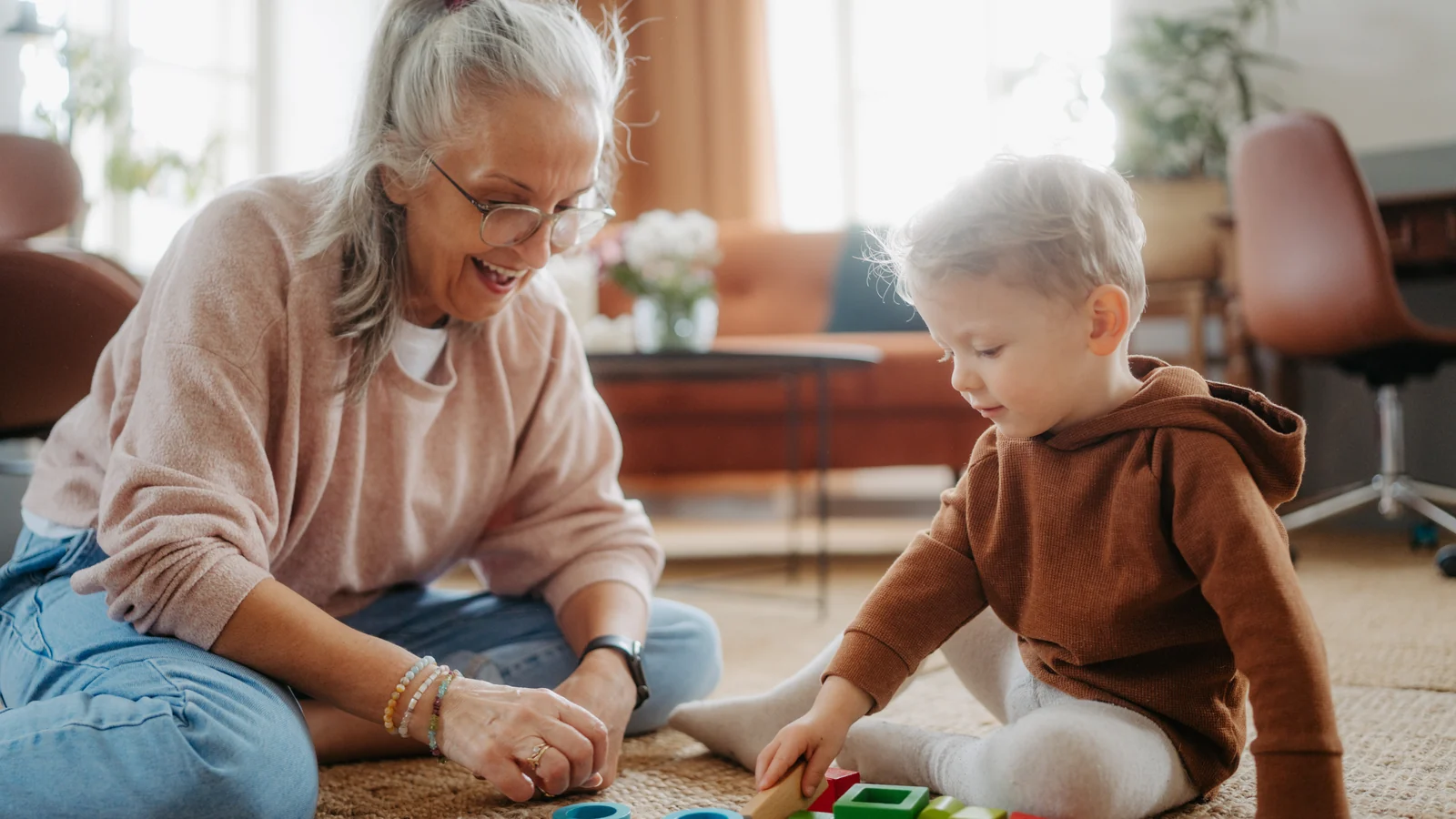 Montessori for grandparents: a grandmother sits on a rug beside her toddler grandchild as he plays with colorful blocks