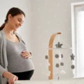 Smiling pregnant woman with a hand on her belly beside a white bassinet in a bright, simple Montessori nursery